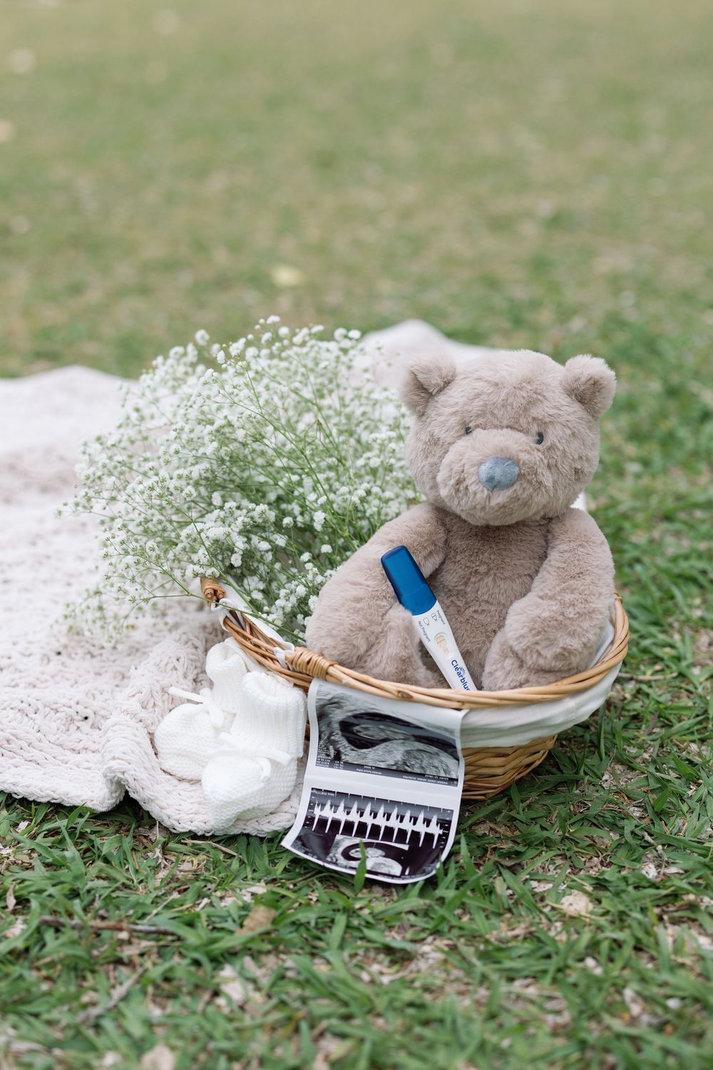 Wicker basket with teddy bear, pregnancy test, ultrasound photos, baby booties, and white flowers on green grass.