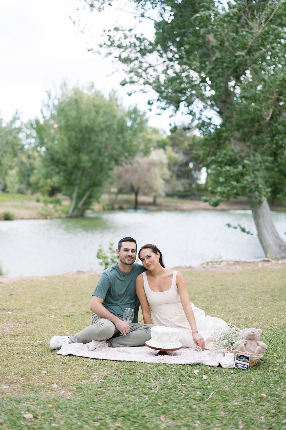 Couple sitting on a blanket by a lake in a park, smiling at the camera on a peaceful outdoor picnic.