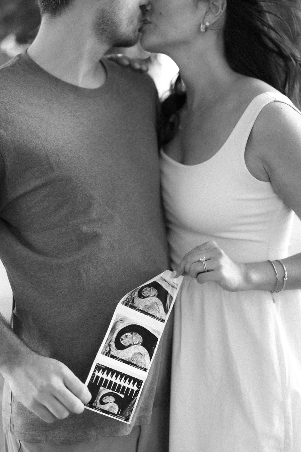 Black and white photo of a couple kissing while holding ultrasound photos, announcing their pregnancy.