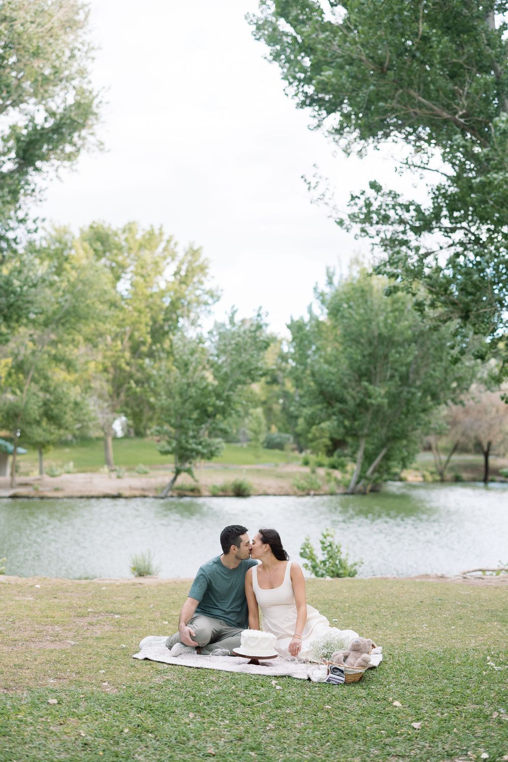 Couple sharing a kiss during a romantic picnic by a serene pond, surrounded by lush green trees in a park.