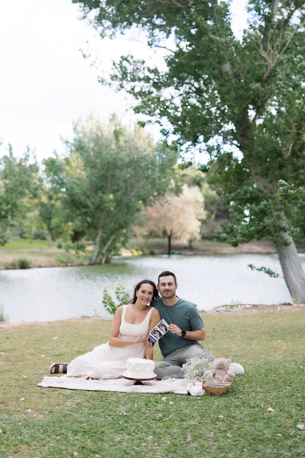 Couple sitting on blanket by lake in park, holding ultrasound photos, announcing pregnancy outdoors.