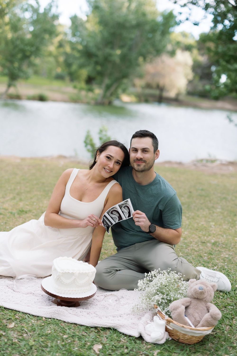 Couple sitting on blanket in park holding ultrasound photos with baby items, cake and teddy bear nearby.