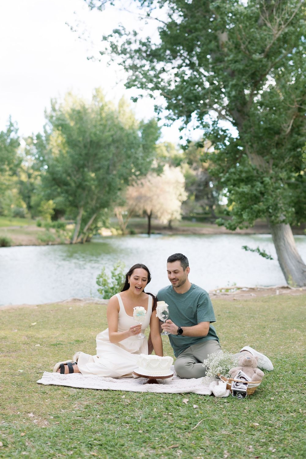 A couple enjoys a romantic lakeside picnic on a blanket, laughing and sharing drinks surrounded by lush green trees.