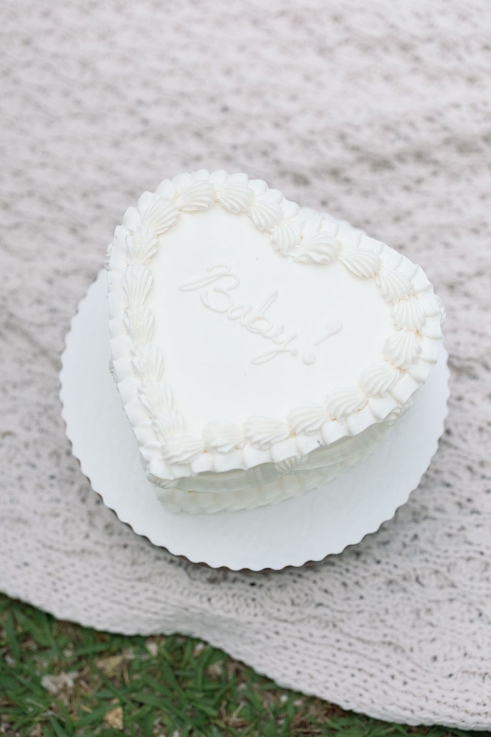 Heart-shaped white cake with Baby written in frosting, displayed on a white plate over a knit blanket outdoors.
