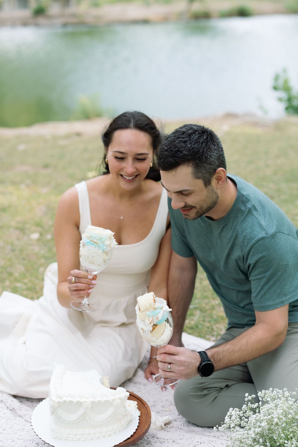 Couple sitting on blanket by lake, smiling while holding wedding cake pops with white tiered cake nearby.