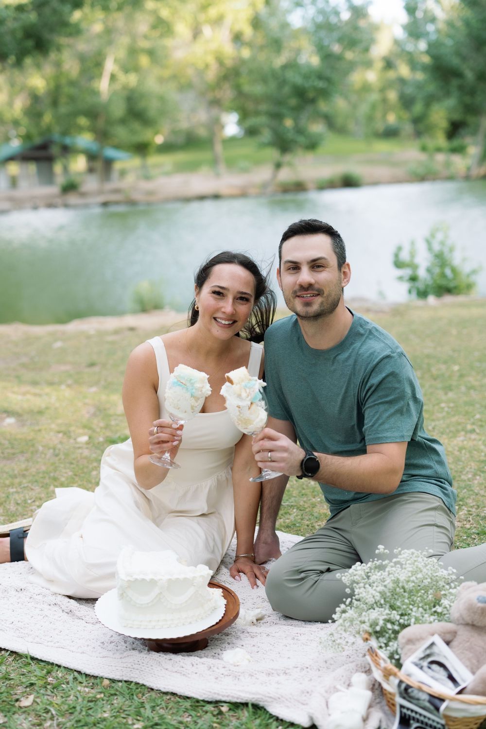 Couple smiling at a lakeside picnic with white cake and cotton candy, sitting on a blanket in a lush green park.