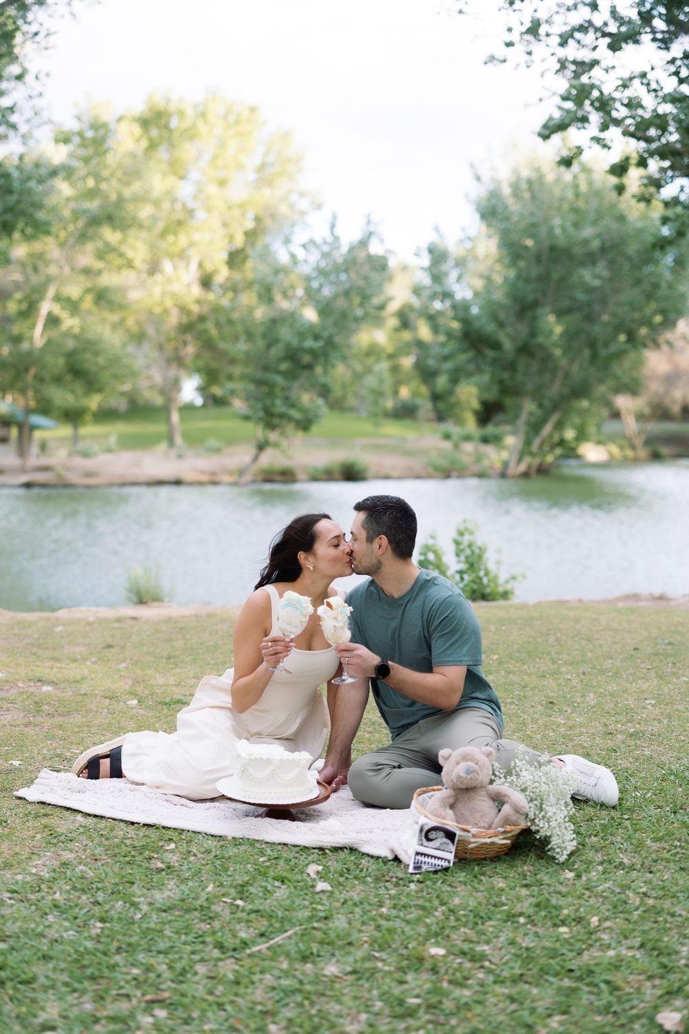Couple sharing a romantic kiss during a picnic by a serene lake in a lush green park on a sunny day.
