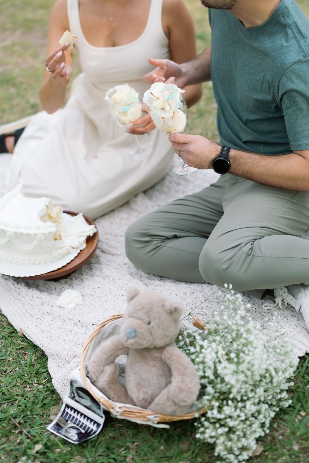 Couple sitting on blanket eating wedding cake with teddy bear, baby's breath flowers, and ultrasound photo at outdoor picnic.