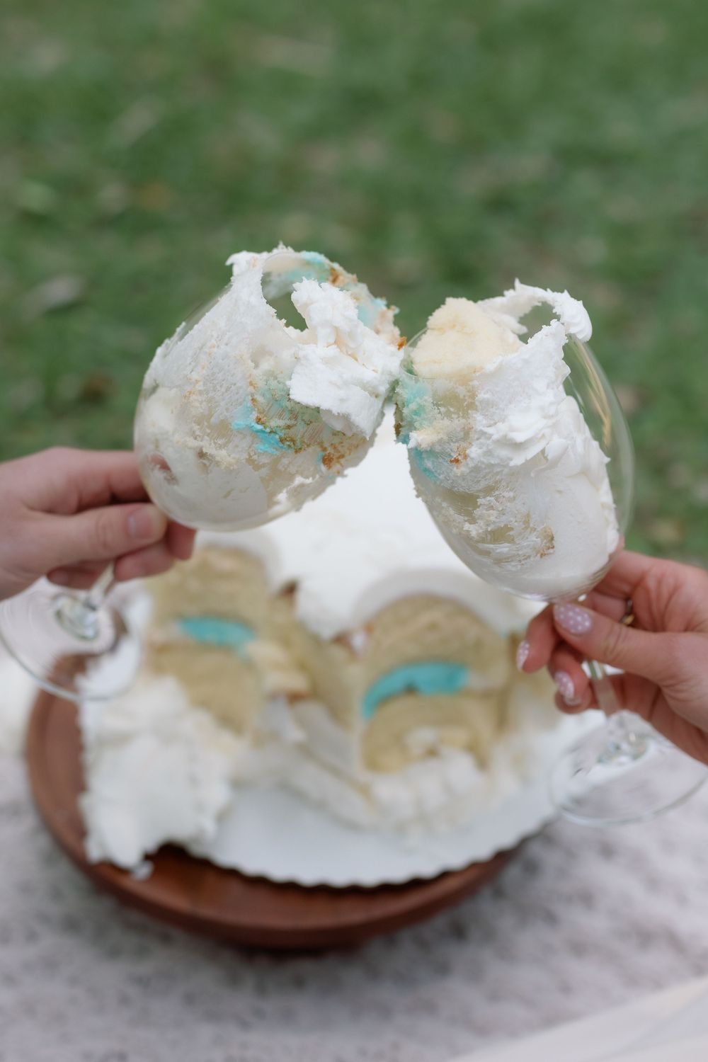 Two hands clinking wine glasses filled with white cake and blue frosting, with a sliced white celebration cake in the background.