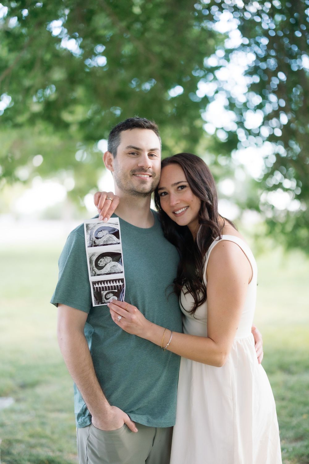 Happy couple holding ultrasound photos outdoors, announcing pregnancy under green trees.