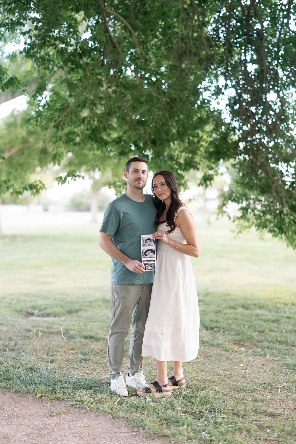 Couple standing under tree in park holding ultrasound photo, man in green shirt and woman in white dress.