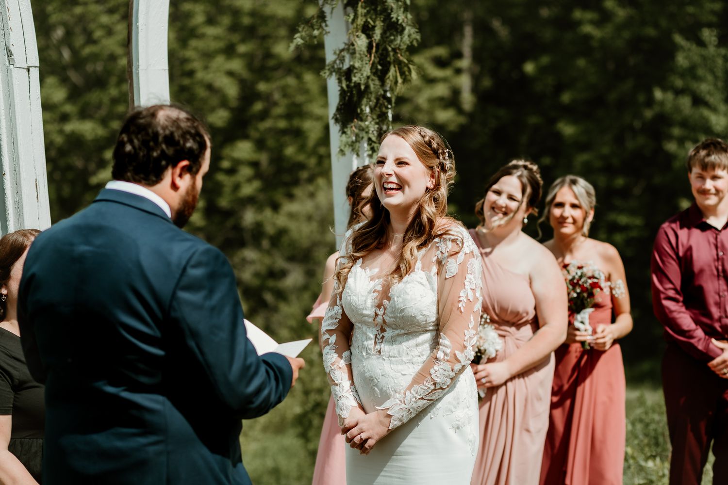Bride laughing joyfully during outdoor wedding ceremony with bridesmaids in dusty rose dresses standing nearby.