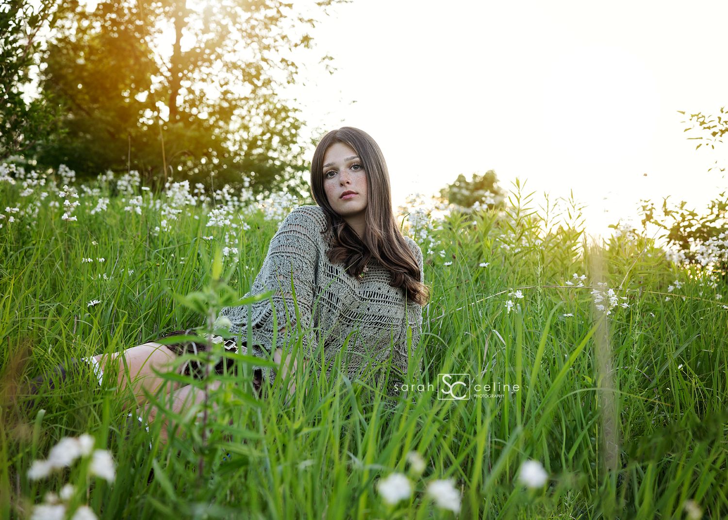 Young woman in a patterned dress sitting in a lush green field with wildflowers during golden hour sunset.