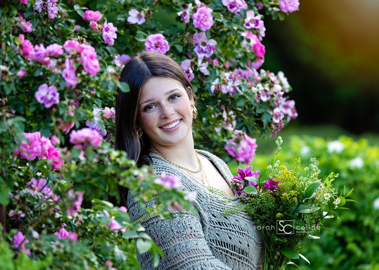 Young woman smiling among blooming pink roses and lush green foliage in a garden setting.