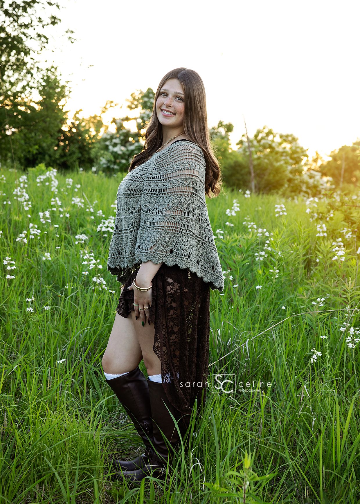 Young woman in lace skirt, knit shawl, and tall boots smiling in a sunlit green meadow with wildflowers.