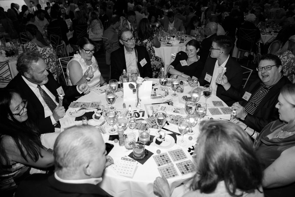 Black and white overhead view of formally dressed guests playing bingo at a large round table during a gala.