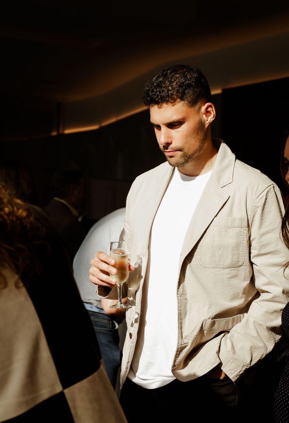 A stylish man in a beige blazer and white tee holds a glass drink at a dimly lit social event.