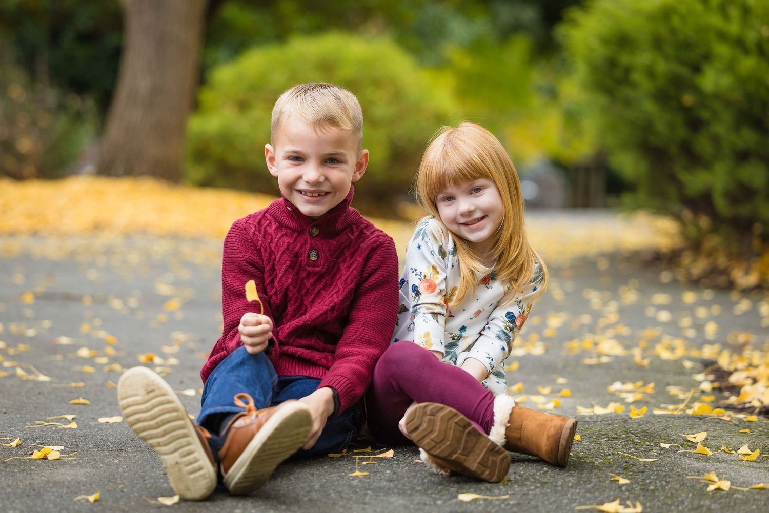 Two young children, a boy and girl, sit smiling on a leaf-covered path in an autumn park.