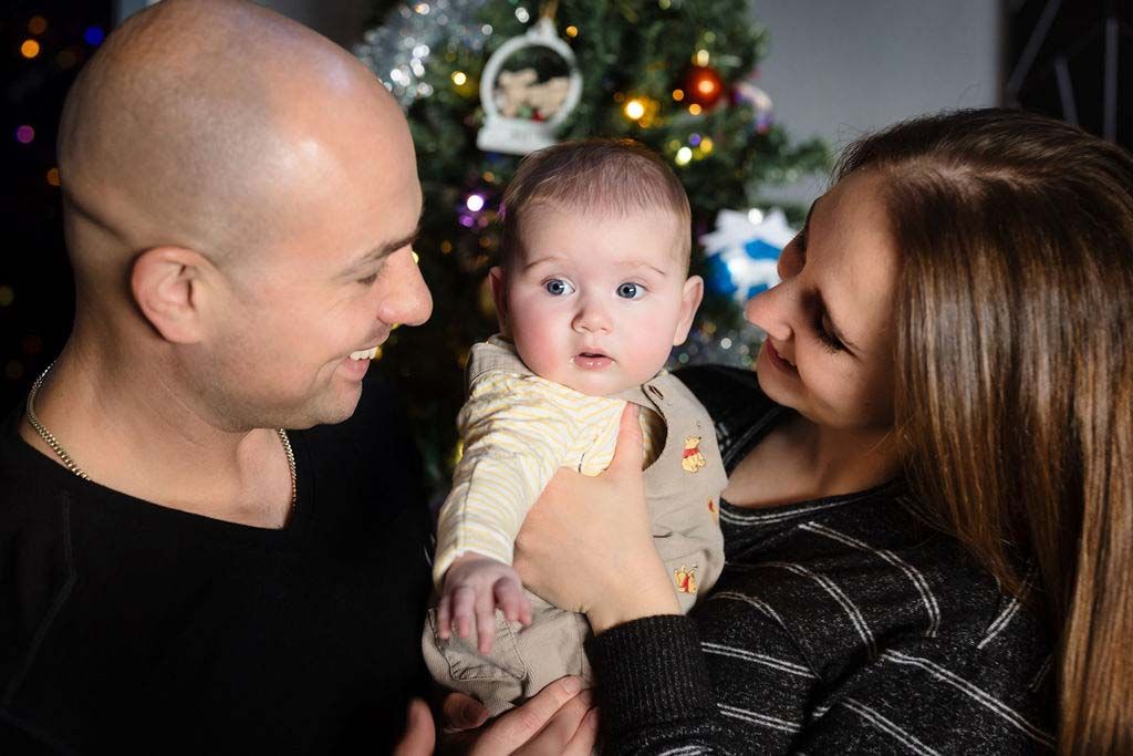 A young family with a bald father and mother holding their wide-eyed baby in front of a decorated Christmas tree.