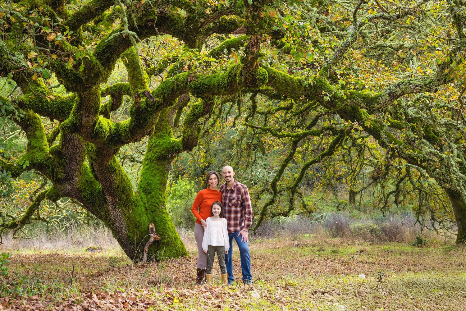 Family of three posing under a massive moss-covered oak tree in an autumn forest setting.