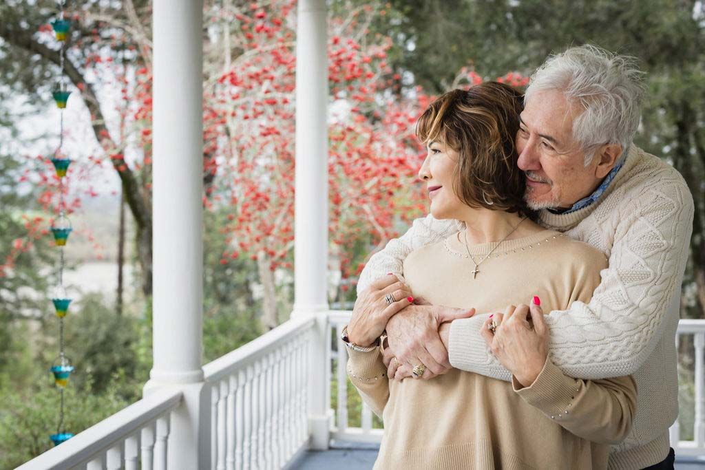 Happy senior couple embracing on a porch, surrounded by autumn trees with red foliage.