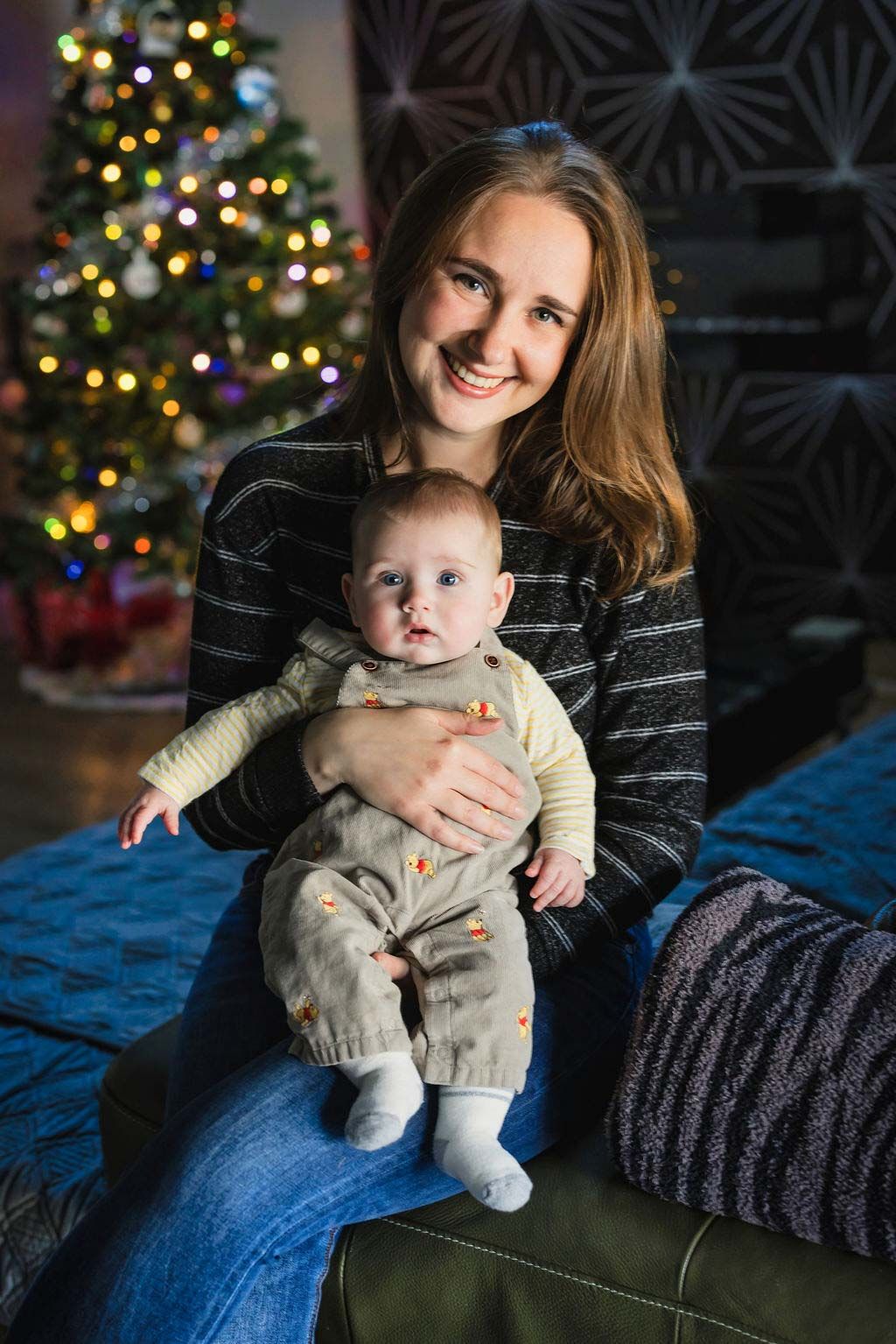 Smiling young mother holding her baby in a knit outfit, sitting in front of a decorated Christmas tree.