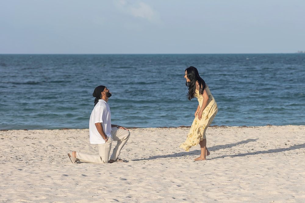 Man kneeling on sandy beach proposing to woman in yellow dress with ocean waves in background.