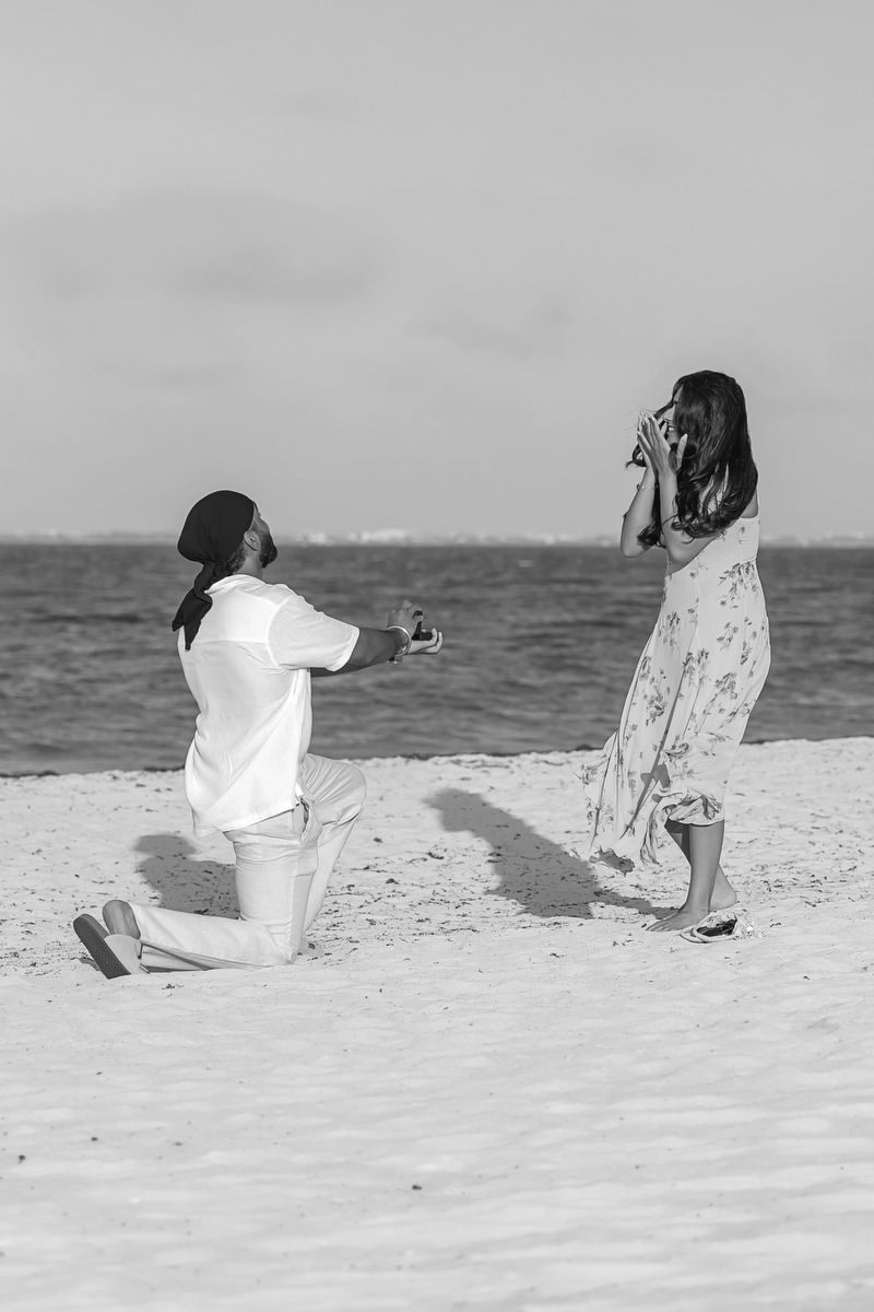 Man proposes on one knee to woman on sandy beach, black and white romantic engagement photo by the ocean.