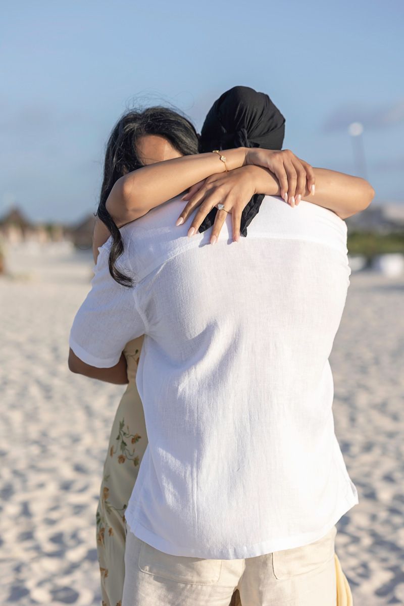 Couple embracing in a warm hug on a white sand beach, woman wearing floral skirt and man in white linen shirt.