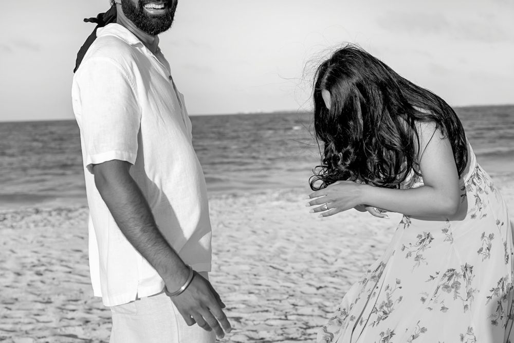 Black and white photo of couple on beach, woman laughing and bending forward in floral skirt beside smiling man.