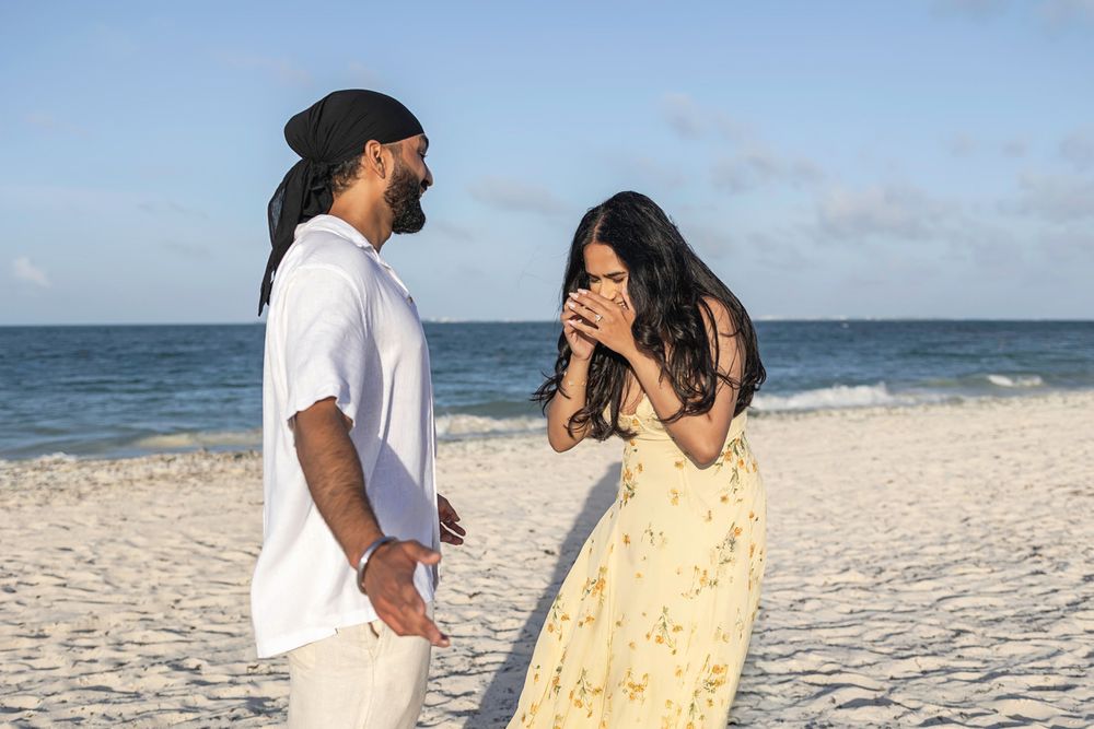 Couple sharing a romantic moment on a white sand beach, woman in yellow floral dress covering her mouth in surprise.