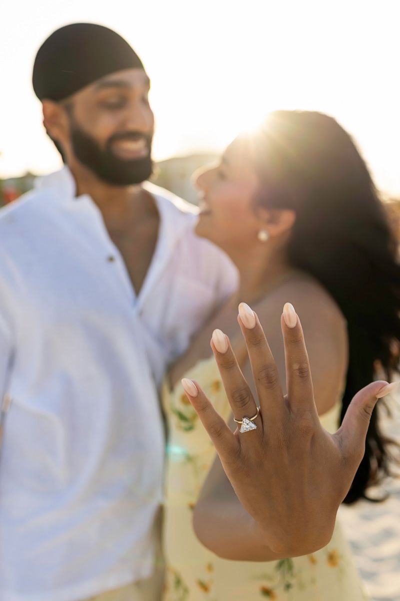 Couple celebrating engagement on beach at sunset, woman showing diamond ring close-up.