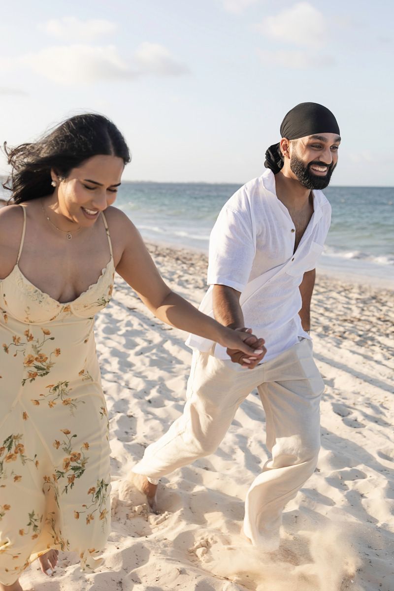 A joyful couple holds hands while running on a sandy beach, the woman in a floral dress and man in white linen.