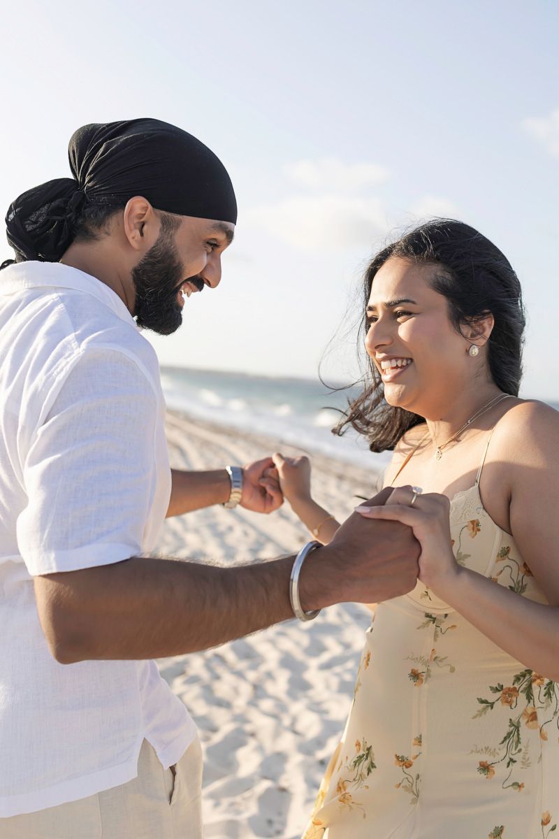 A joyful couple holds hands and smiles at each other on a sunny beach, man wearing turban and woman in floral dress.