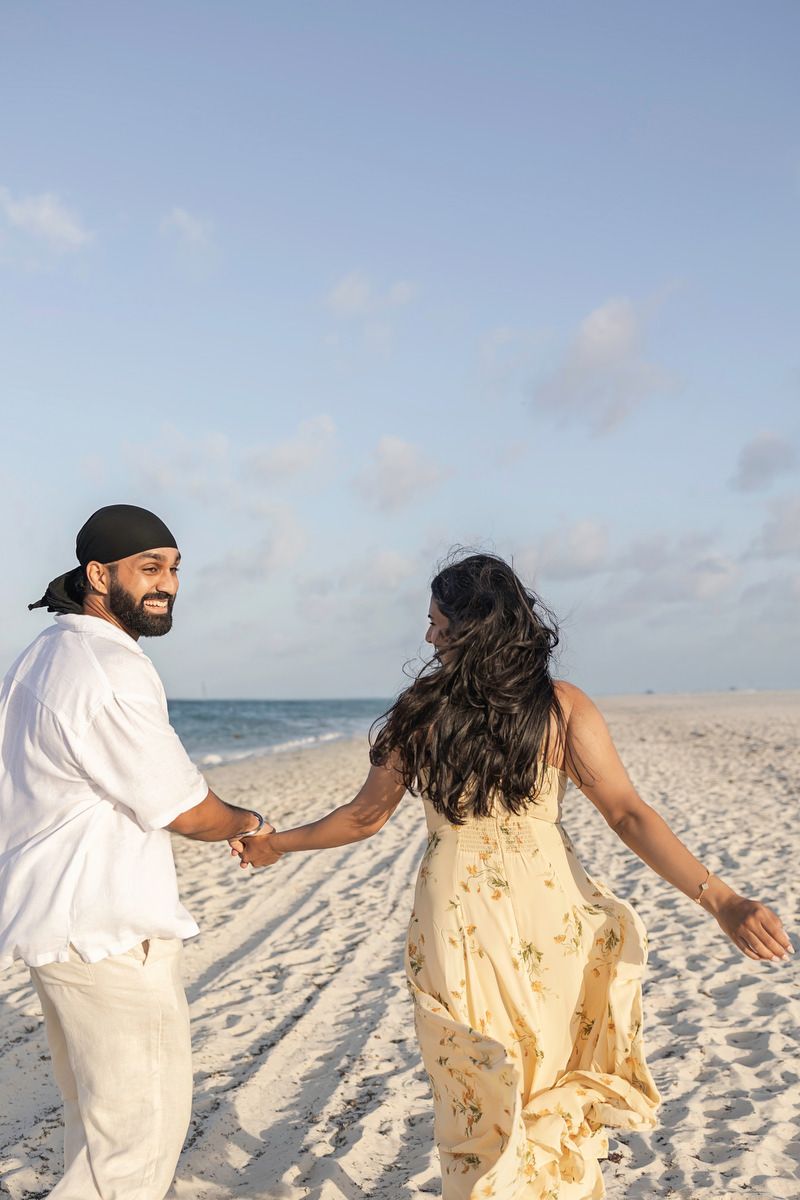 Happy couple holding hands walking on white sandy beach with ocean backdrop.