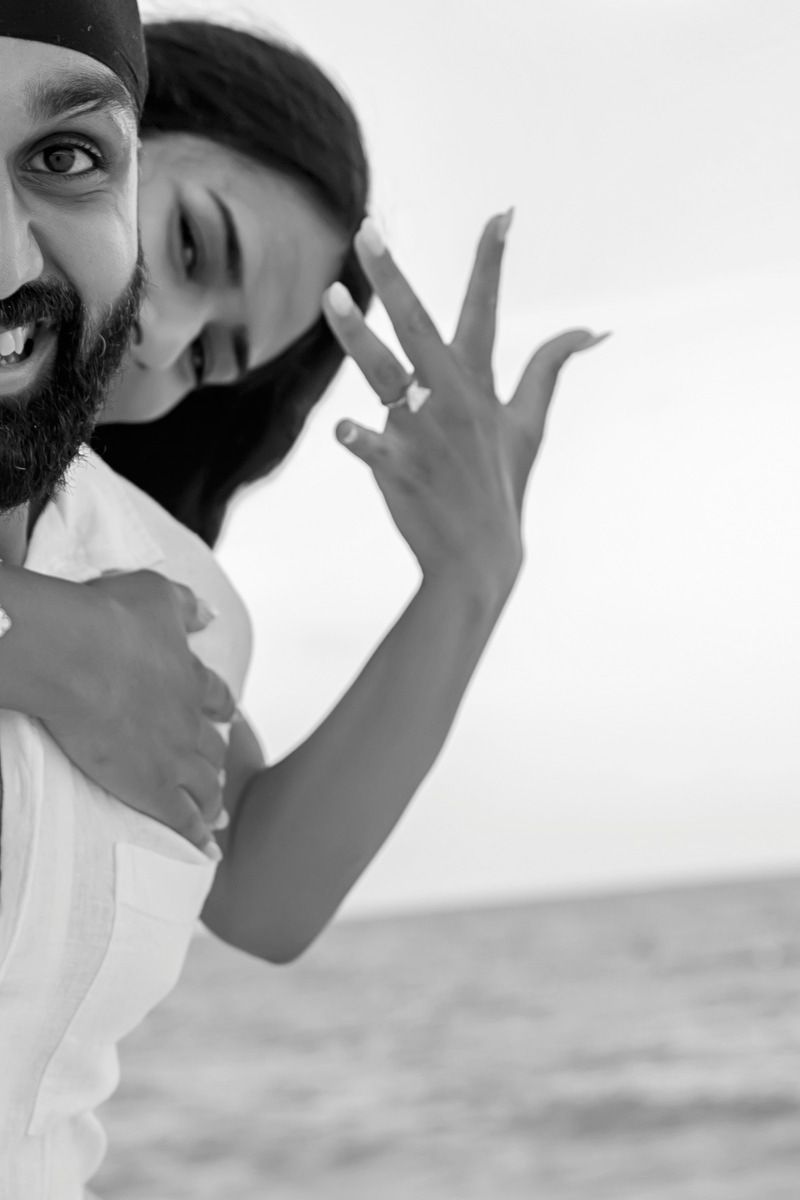 A happy couple poses at the beach in black and white, the woman showing off her engagement ring while hugging her partner.