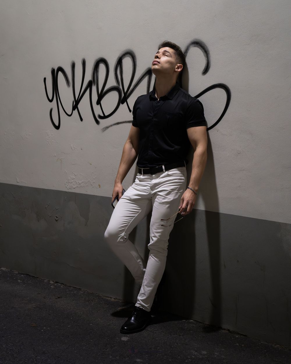 A muscular man in black polo and white pants leans against a graffiti wall, head tilted back, wearing black loafers.