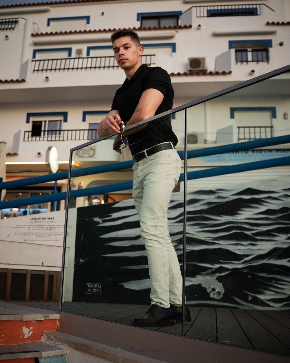 Young man in black shirt and light pants posing on glass railing terrace with ocean mural backdrop.