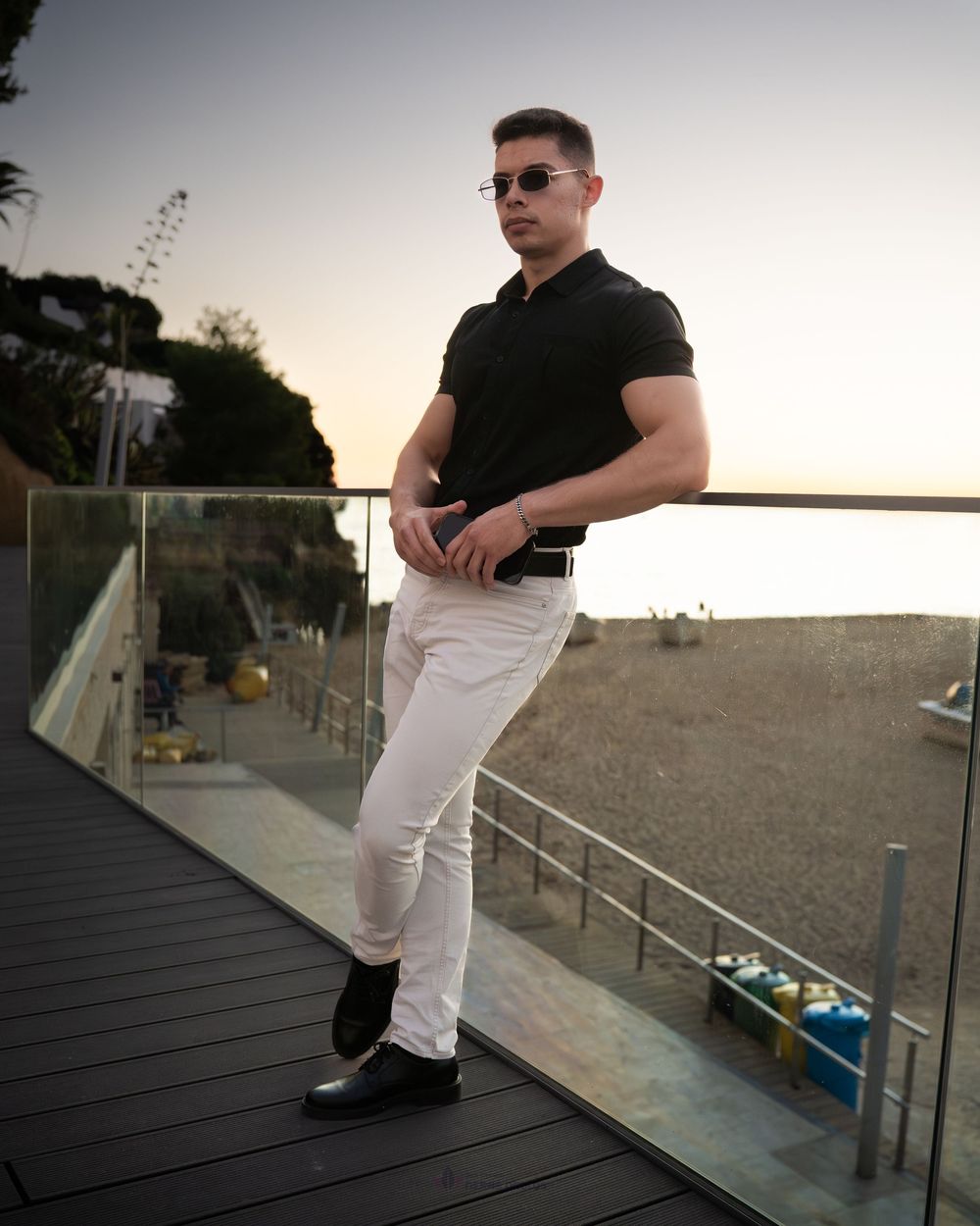 Stylish man in black polo and white jeans posing on a glass-railed balcony overlooking a beach at dusk.