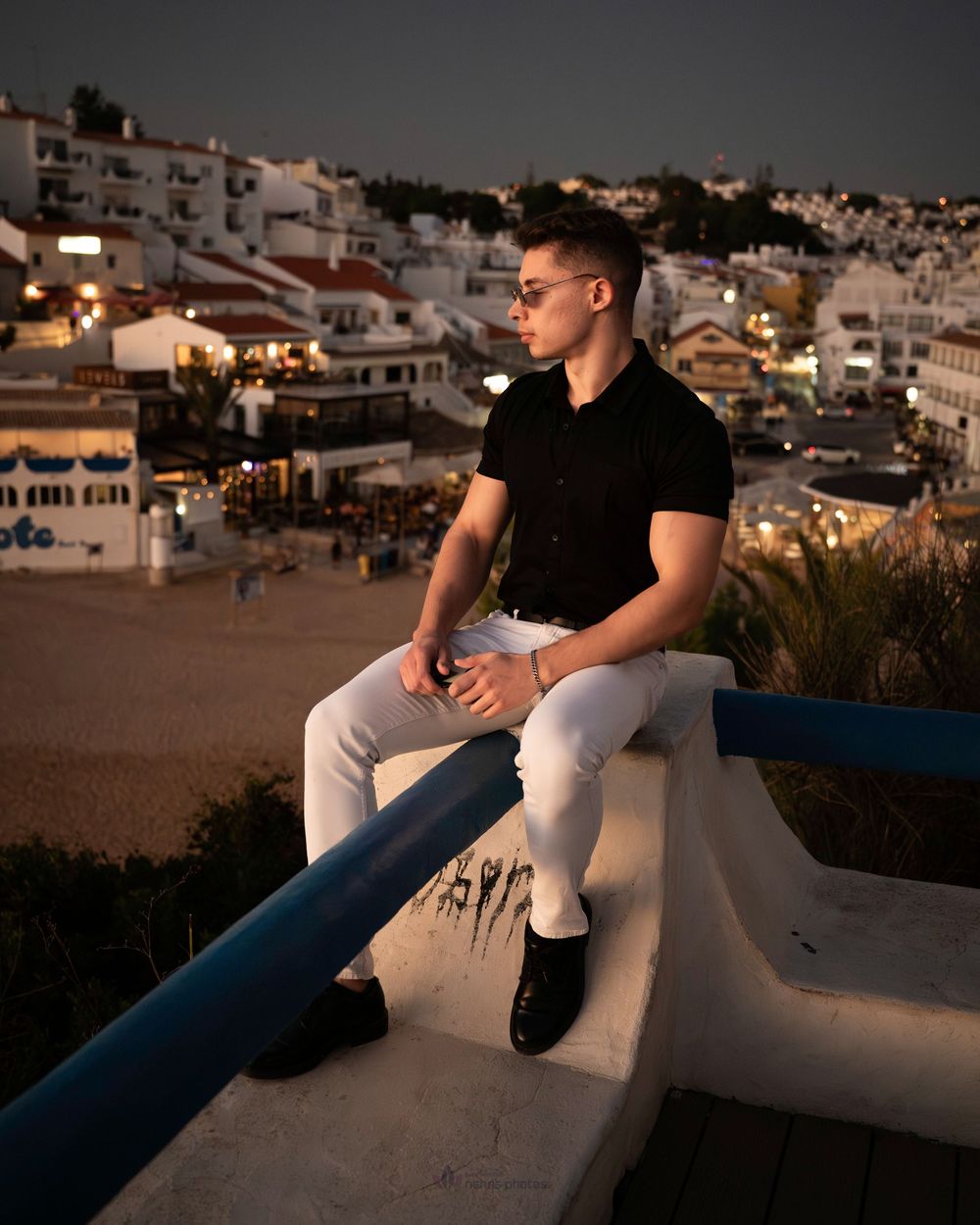 Young man in black shirt and white jeans sitting on a wall at dusk overlooking a Mediterranean coastal town.