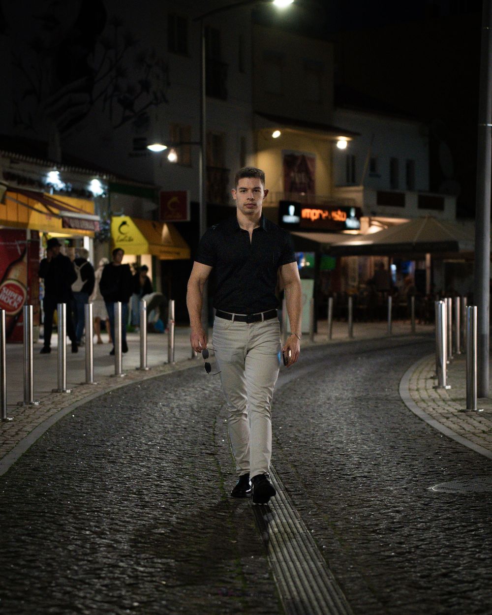 Man in black shirt and gray pants walking on cobblestone street at night with lit shops in background.