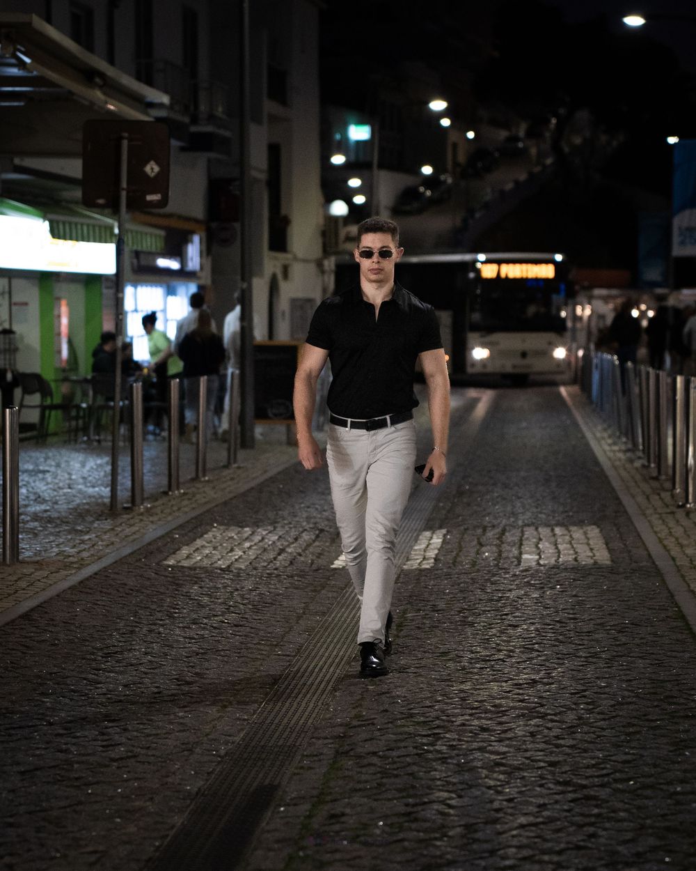 Stylish man in black polo and gray pants walking confidently on cobblestone street at night.