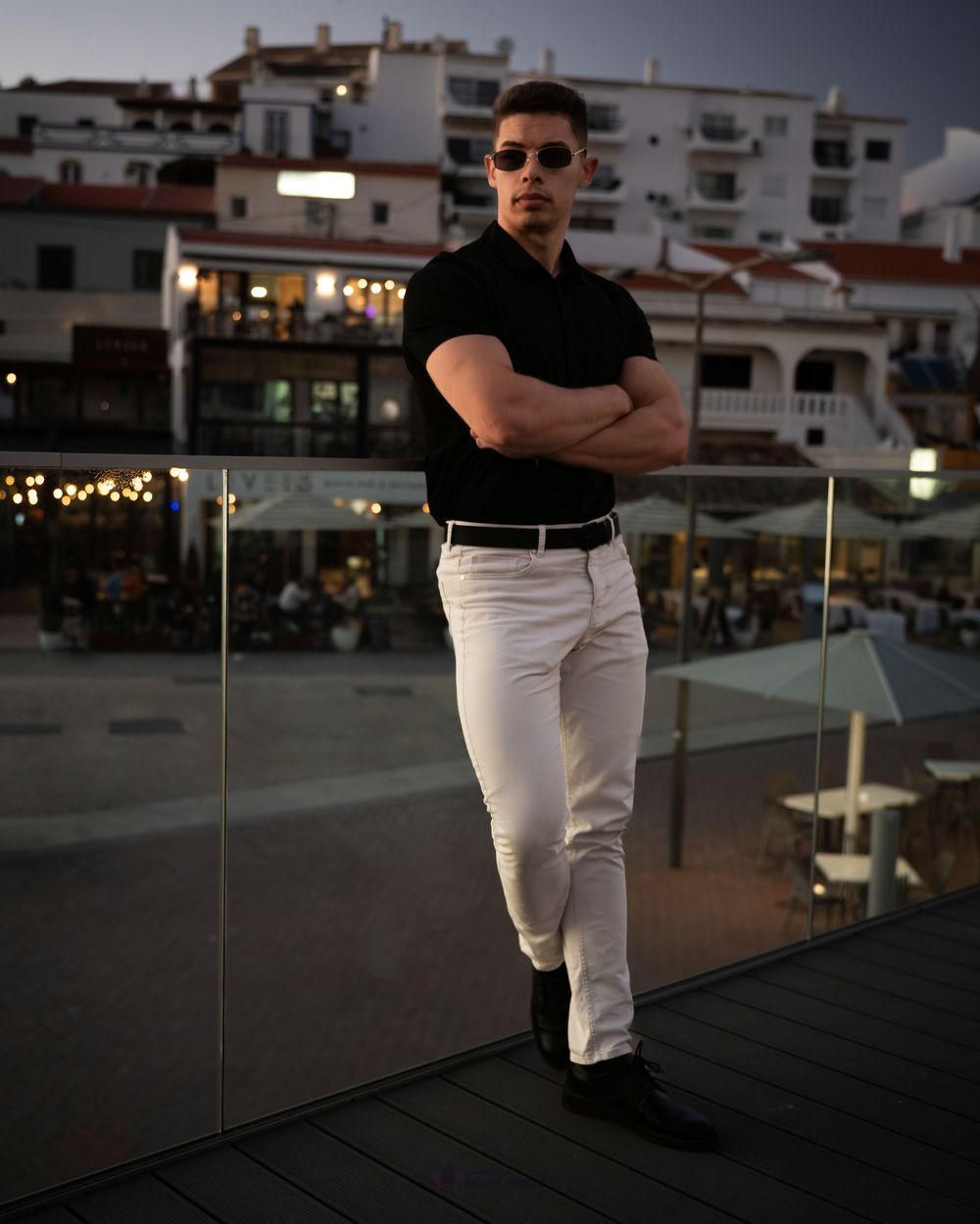 Man in black polo and white pants poses with arms crossed on a rooftop terrace at dusk.