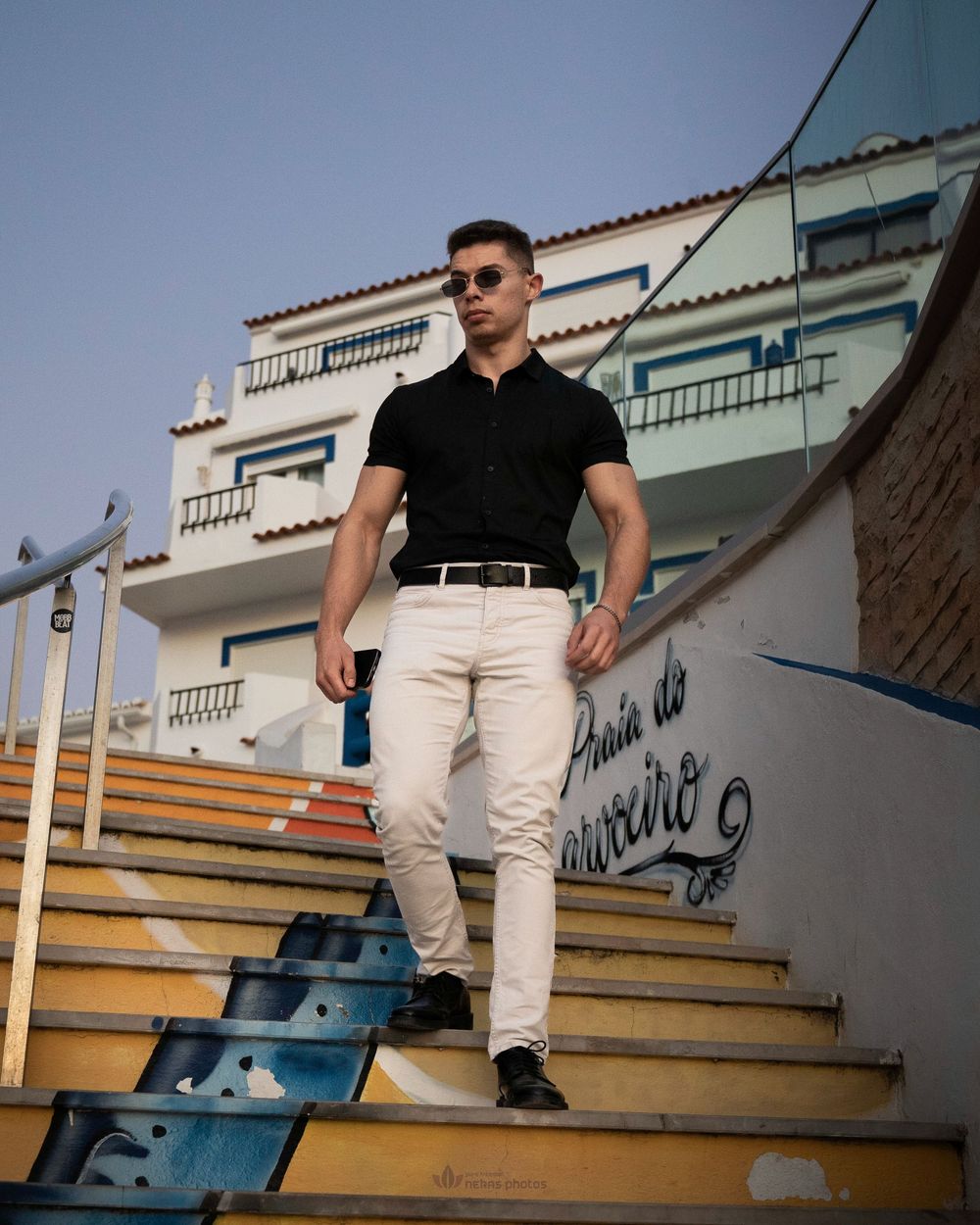 Muscular man in black shirt and white pants walking down colorful stairs with graffiti near a white building.