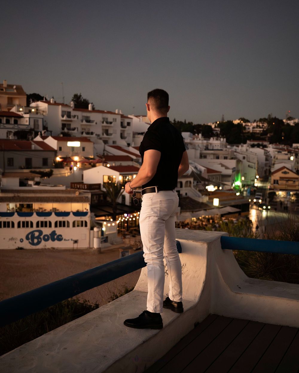 Man in black shirt and white jeans standing on a terrace overlooking a lit Mediterranean town at dusk.