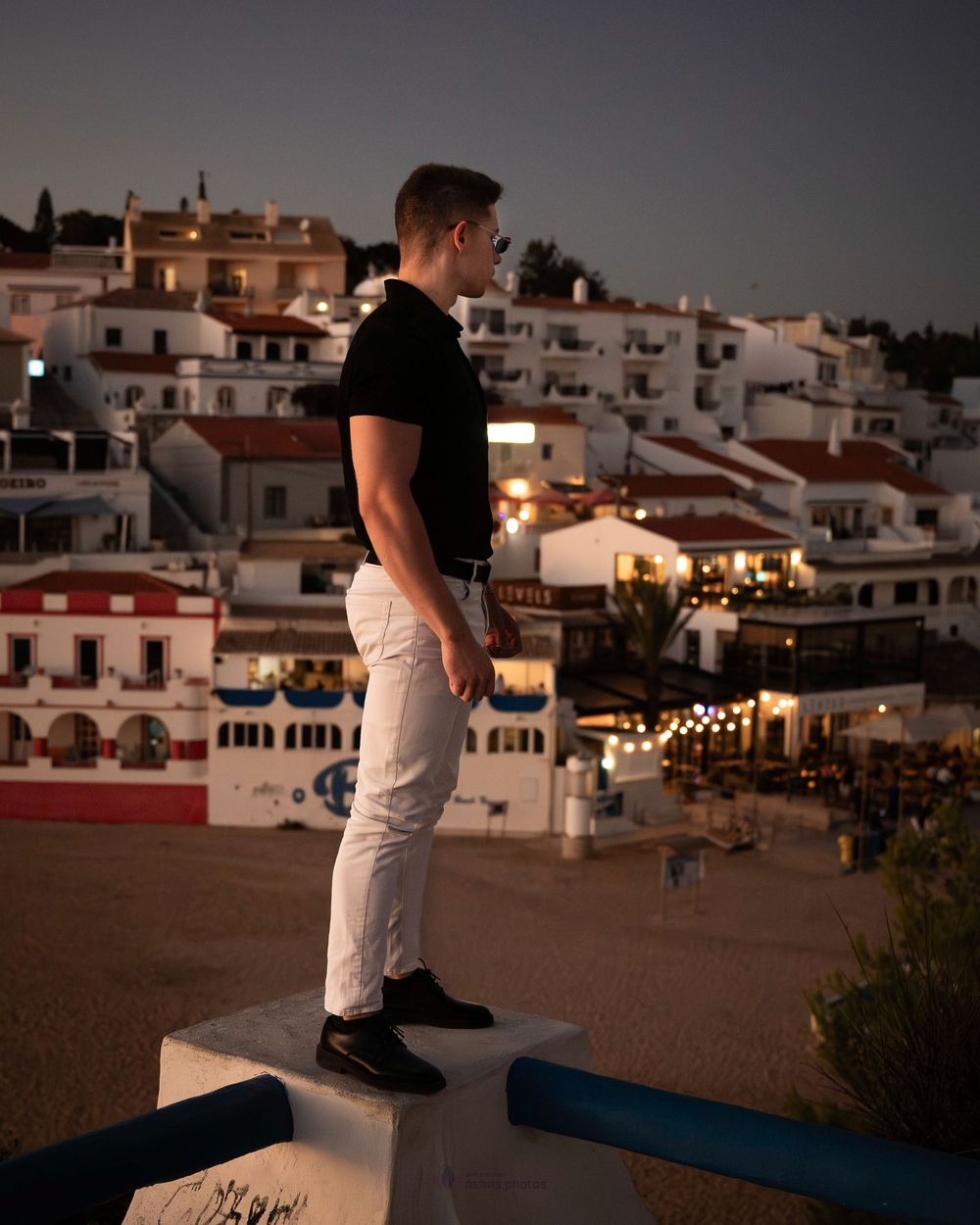 Man in white pants and black shirt standing on a pedestal overlooking a lit Mediterranean village at dusk.