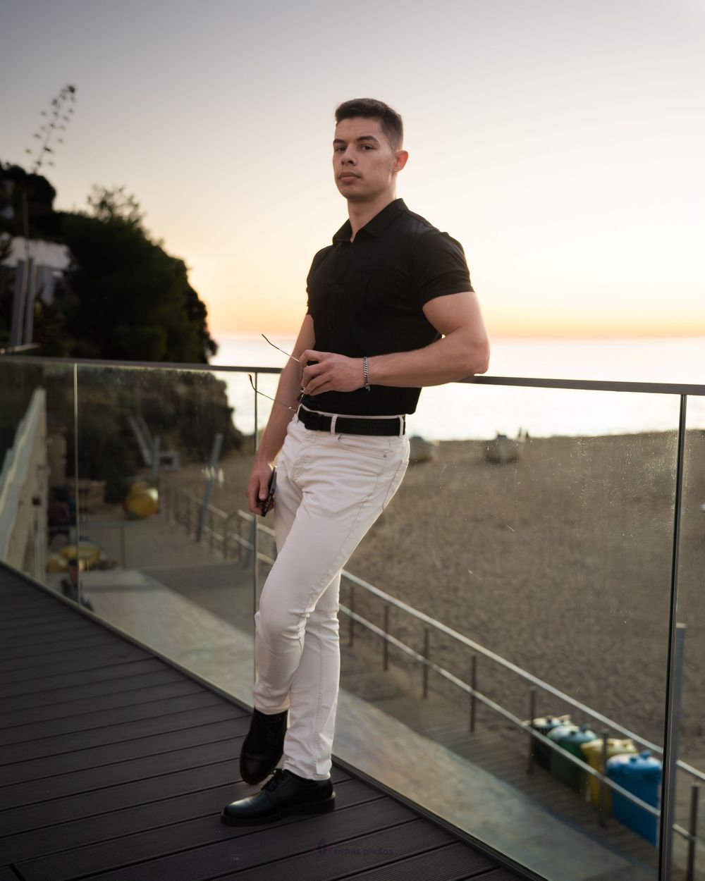 Young man in black polo and white pants poses on a rooftop deck overlooking a beach at sunset.