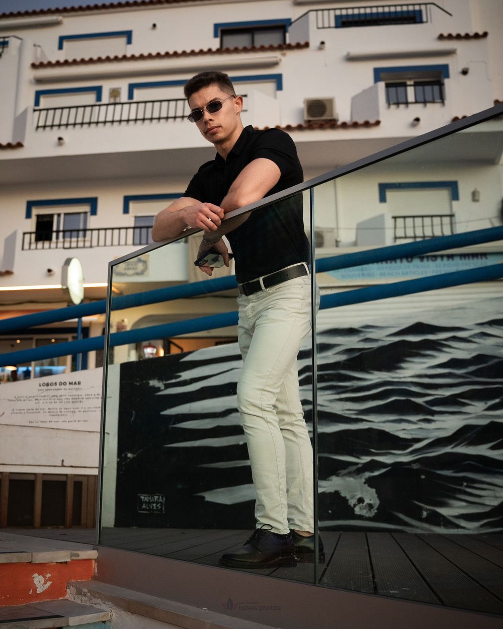 Man in black shirt and white jeans leaning on glass railing with coastal building backdrop.
