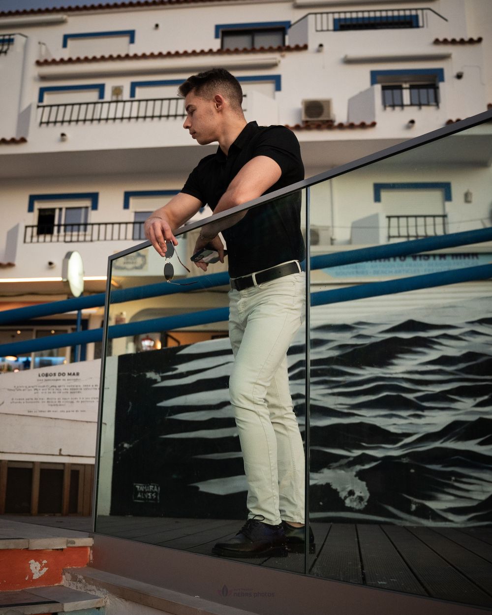 Man in black shirt and white jeans leaning on glass railing with ocean mural and white building backdrop.