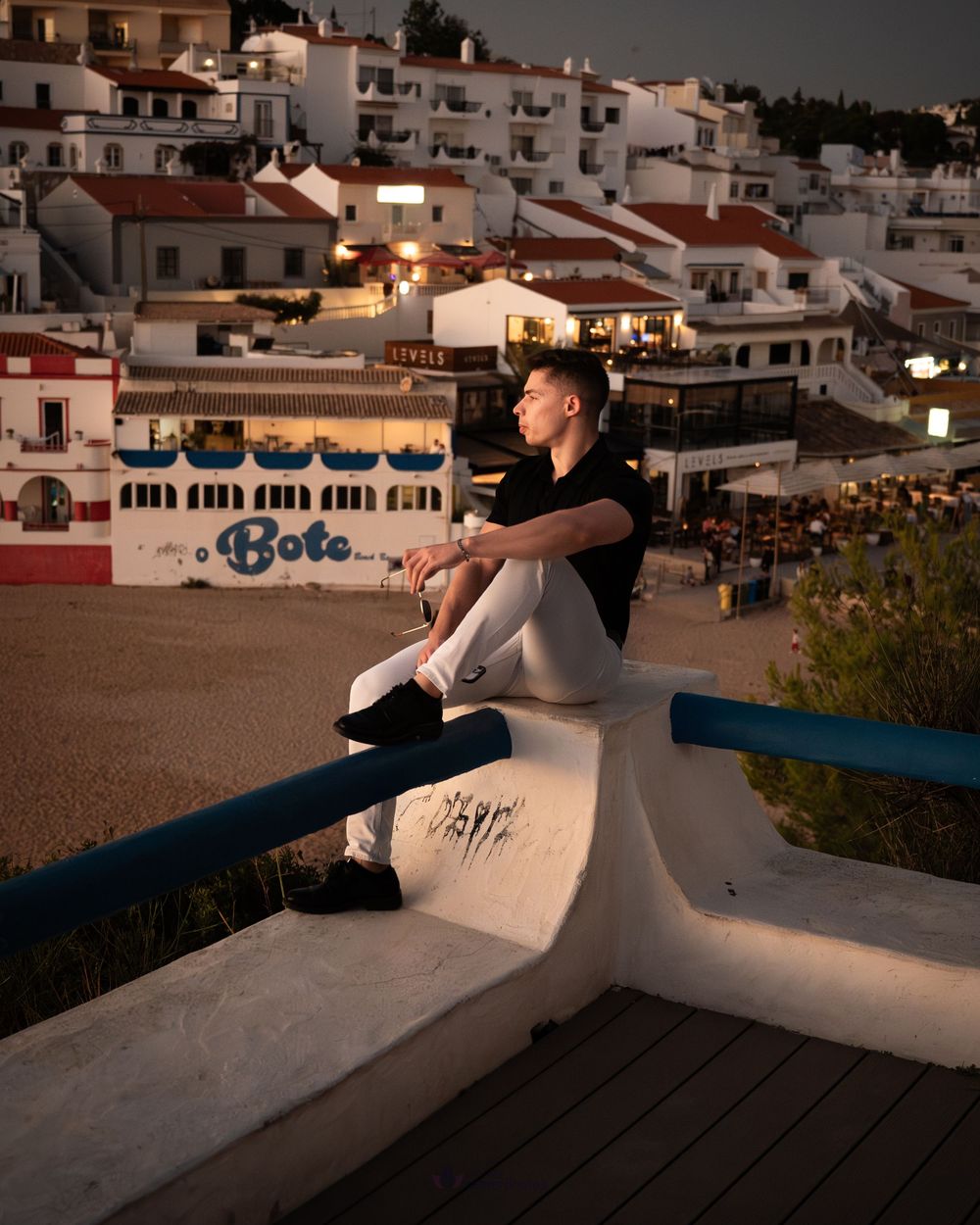 Young man sitting on a white wall at dusk overlooking a picturesque Mediterranean coastal village with white buildings.